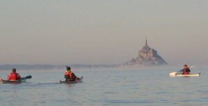 Trois kayakistes naviguant en mer dans la brume matinale avec le Mont Saint-Michel en toile de fond.